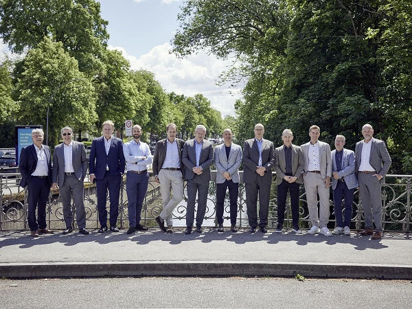 Eine Gruppe von zwölf Männern in Anzügen steht auf einer Brücke, umgeben von Bäumen und einem klaren Himmel im Hintergrund. Sie sind in einem entspannten, freundlichen Arrangement aufgestellt.