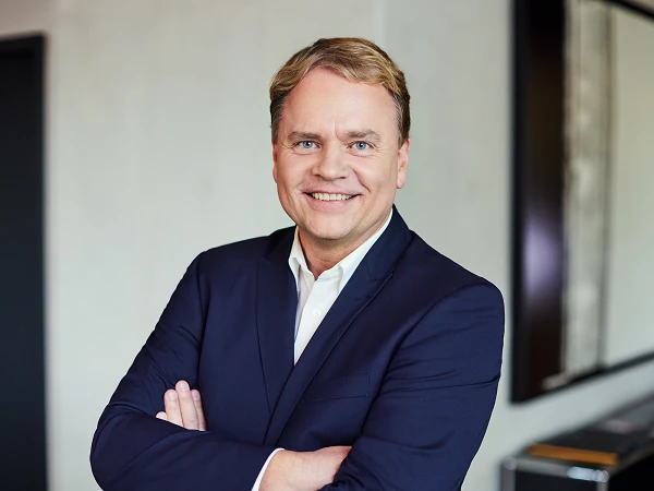Ein gut gekleideter Mann mit blonden, kurzen Haaren lächelt freundlich in die Kamera, während er mit verschränkten Armen vor einem hellen, modernen Hintergrund steht.