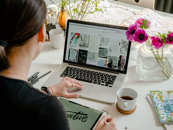 Eine Person sitzt an einem Tisch und arbeitet an einem Laptop, während sie auf einem Tablet mit einer künstlerischen Handschrift "creative" schreibt. Auf dem Laptop-Bildschirm sind verschiedene Designideen zu sehen. Neben dem Laptop stehen eine Tasse Kaffee und eine Vase mit frischen Blumen.