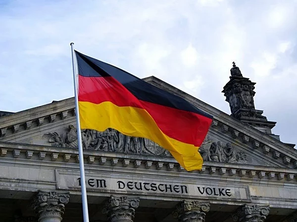 Die deutsche Flagge in Schwarz-Rot-Gelb weht vor dem Reichstagsgebäude in Berlin, das mit einem kunstvollen Bauwerk und der Inschrift „Dem Deutschen Volke“ verziert ist.