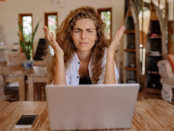 Eine frustrierte Frau mit lockigem, braunem Haar sitzt an einem Holztisch vor einem Laptop und hebt die Hände in die Luft, als Zeichen der Verwirrung oder Aufregung. Im Hintergrund sind minimalistisch eingerichtete Möbel und Pflanzen sichtbar.