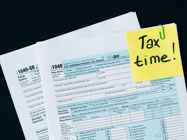 Auf einem schwarzen Tisch liegen zwei Formulare „1040-SR“ zur Einkommenssteuererklärung, und ein gelbes Haftnotizpapier mit dem Text „Tax time!“ ist mit einer Büroklammer befestigt.