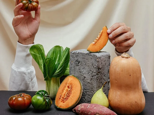 Eine Hand hält eine Tomate, während die andere ein Stück Melone präsentiert; verschiedene frische Gemüse und Obst, darunter Pak Choi, Süßkartoffel, Birne und Kürbis, sind um einen grauen Block angeordnet.