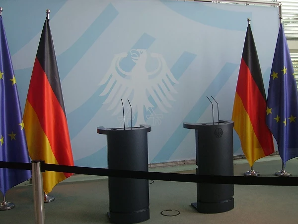 Zwei Podeste mit Mikrofonen stehen vor einem Hintergrund mit dem deutschen Bundesadler und einer Reihe von Flaggen: Deutschland, Europa und der EU. Ein schwarzes Absperrband trennt den Bereich sichtbar ab.