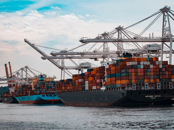 Containerfrachtsschiffe laden und entladen ihre bunten Container in einem Hafen, während Kräne über ihnen stehen und am Himmel Wolken zu sehen sind.