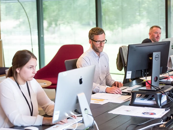 Drei Personen arbeiten konzentriert an Schreibtischen in einem modernen Büroraum, umgeben von großen Fenstern und einer gemütlichen Sitzecke.