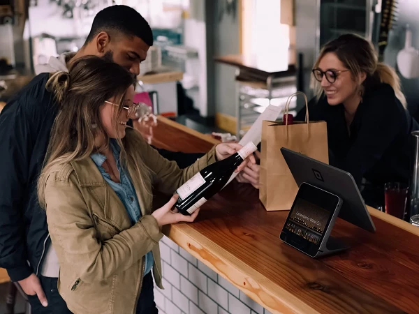Eine Frau hält eine Flasche Wein und zeigt sie einem Mann, während eine Bartenderin an einem Tresen hinter ihnen mit einem Tablet und einer Papiertüte steht.