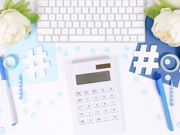 Ein Schreibtisch mit einer weißen Tastatur, einem Taschenrechner und verschiedenen Büromaterialien, darunter eine blaue und eine durchsichtige Klebebandrolle, eine blaue Stift und Papierschnipsel in Form von Hashtags, umgeben von weißen Blumen und blauen Punkten.