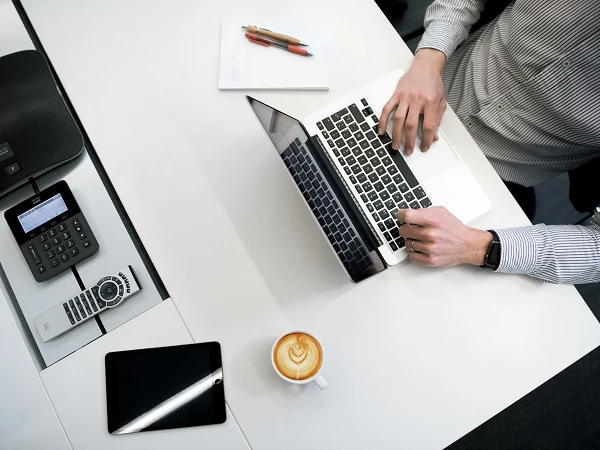 Eine Person tippt auf einem Laptop, während auf dem Tisch ein Smartphone, ein Tablet und eine Tasse mit Kaffee stehen. Im Hintergrund sind Schreibutensilien und ein Notizblock zu sehen.