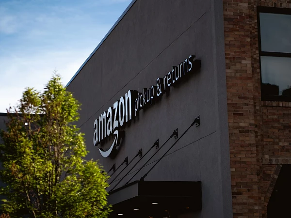 Das Gebäude zeigt ein Schild mit der Aufschrift „amazon pickup & returns“, umgeben von einem Baum und einem klaren, blauen Himmel.