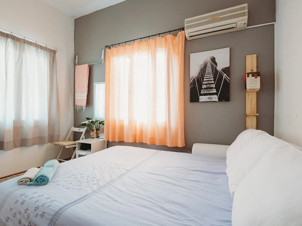 Das Bild zeigt ein gemütliches Schlafzimmer mit einem großen Bett in der Mitte, auf dem eine helle Bettdecke liegt. An der Wand hängt ein schwarz-weißes Bild einer Treppe. Vor einem Fenster mit orangefarbenen Vorhängen steht ein kleiner Tisch mit Pflanzen. Ein Handtuch und eine Decke sind ordentlich auf dem Bett platziert.