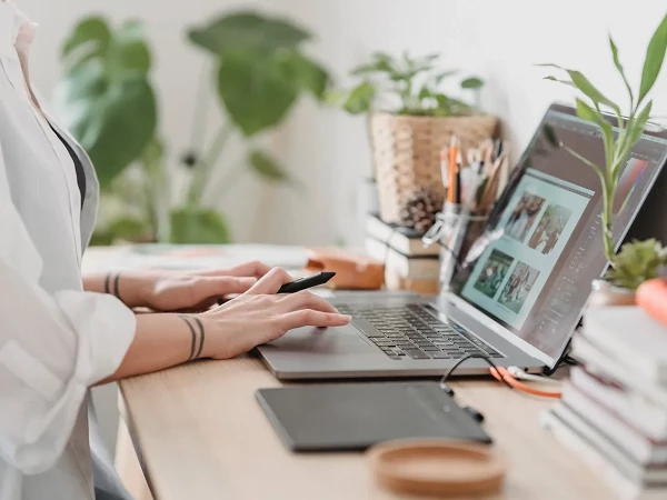 Eine Person sitzt an einem Schreibtisch und arbeitet am Laptop, während sie ein Smartphone in der Hand hält; im Hintergrund sind Pflanzen, Notizbücher und Bürobedarf zu sehen.