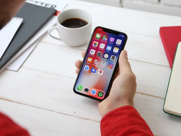Eine Person hält ein Smartphone in der Hand, das auf dem Bildschirm eine Vielzahl von Apps zeigt. Auf dem Tisch liegen zusätzlich ein Notizbuch, ein Stift und eine Tasse Kaffee.
