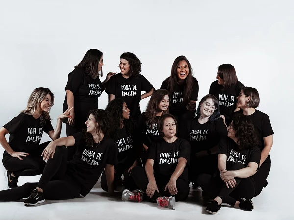 Eine Gruppe von Frauen in schwarzen T-Shirts mit der Aufschrift "Dona de Mim" posiert fröhlich gemeinsam vor einem weißen Hintergrund. Einige Frauen sitzen, andere stehen, und sie lachen und interagieren miteinander.