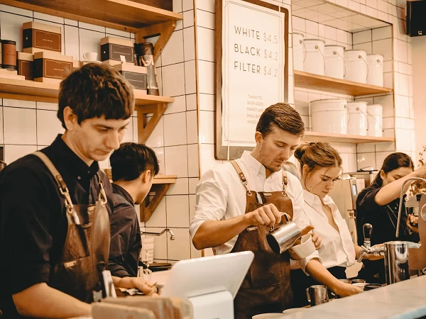 Ein Café mit mehreren Baristas, die an einer Theke arbeiten. Ein Barista gießt Milch in einen Becher, während die anderen Beschäftigten an der Kasse und an der Kaffeemaschine beschäftigt sind. Im Hintergrund sind Regale mit Kaffeeverpackungen und Utensilien zu sehen.