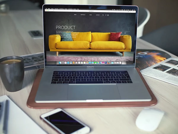 Ein Laptop mit einem geöffneten Bildschirm, der eine Website mit einem gelben Sofa als Hauptmotiv zeigt, steht auf einem Holzschreibtisch. Neben dem Laptop befinden sich eine Tasse Kaffee, ein Notizbuch, ein Stylus, ein Smartphone und eine Zeitschrift.