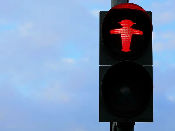 Ein rotes Ampelsignal zeigt eine stilisierte Figur in menschlicher Gestalt mit Hut, während sich darüber ein schwarzes, inaktives Licht befindet und der Himmel teilweise bewölkt ist.