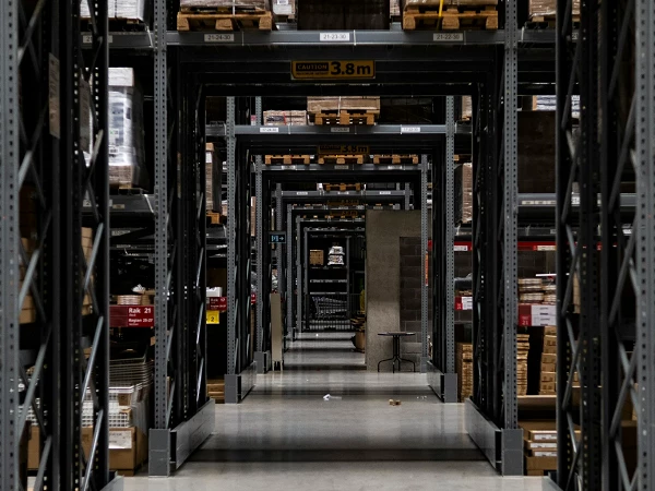 Ein Blick in ein großes Lager mit mehreren Regalen, die gefüllt sind mit Paletten und Kartons. Die Regale sind aus Metall und in gleichmäßigen Abständen angeordnet, wodurch ein perspektivischer Effekt entsteht. Am Ende des Gangbereichs sind einige größere Objekte sichtbar.