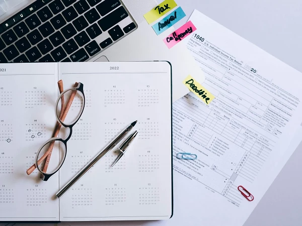 Ein Kalender für die Jahre 2021 und 2022 liegt offen auf einem Tisch, daneben liegen eine Brille, ein Stift und ein ausgefülltes Steuerformular mit bunter Haftnotizen darauf.