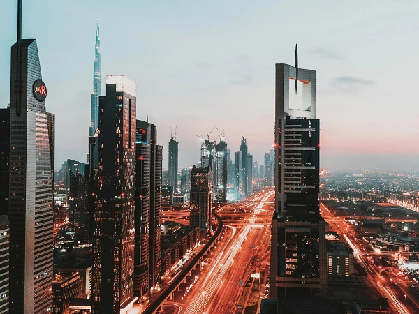 Die Skyline einer modernen Stadt bei Dämmerung ist zu sehen, mit hohen Wolkenkratzern, darunter der Burj Khalifa. Die Straßen sind beleuchtet und zeigen einen Verkehrsfluss, während der Himmel in sanften Blau- und Orangetönen leuchtet.