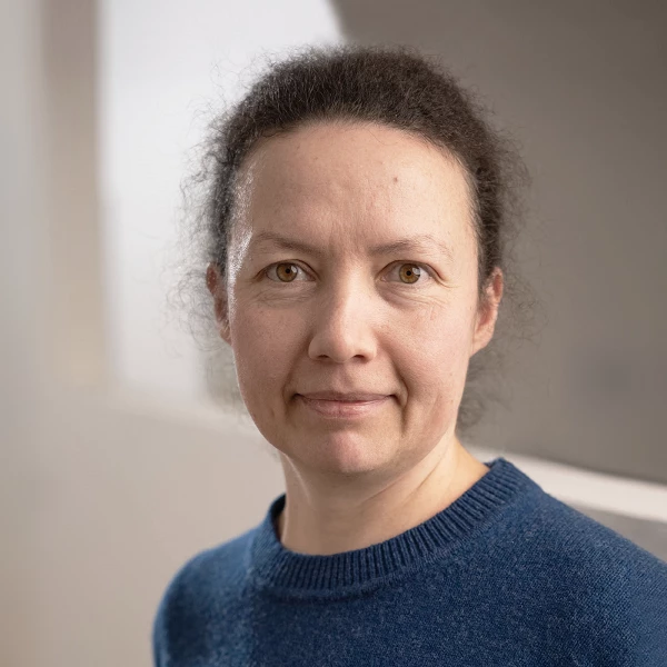 Frau mit lockigem Haar und blauem Pullover in neutraler Büroumgebung, direkter Blick