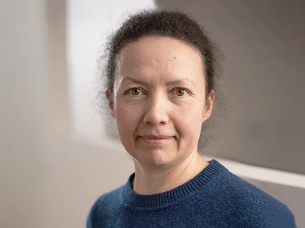 Frau mit lockigem Haar und blauem Pullover in neutraler Büroumgebung, direkter Blick